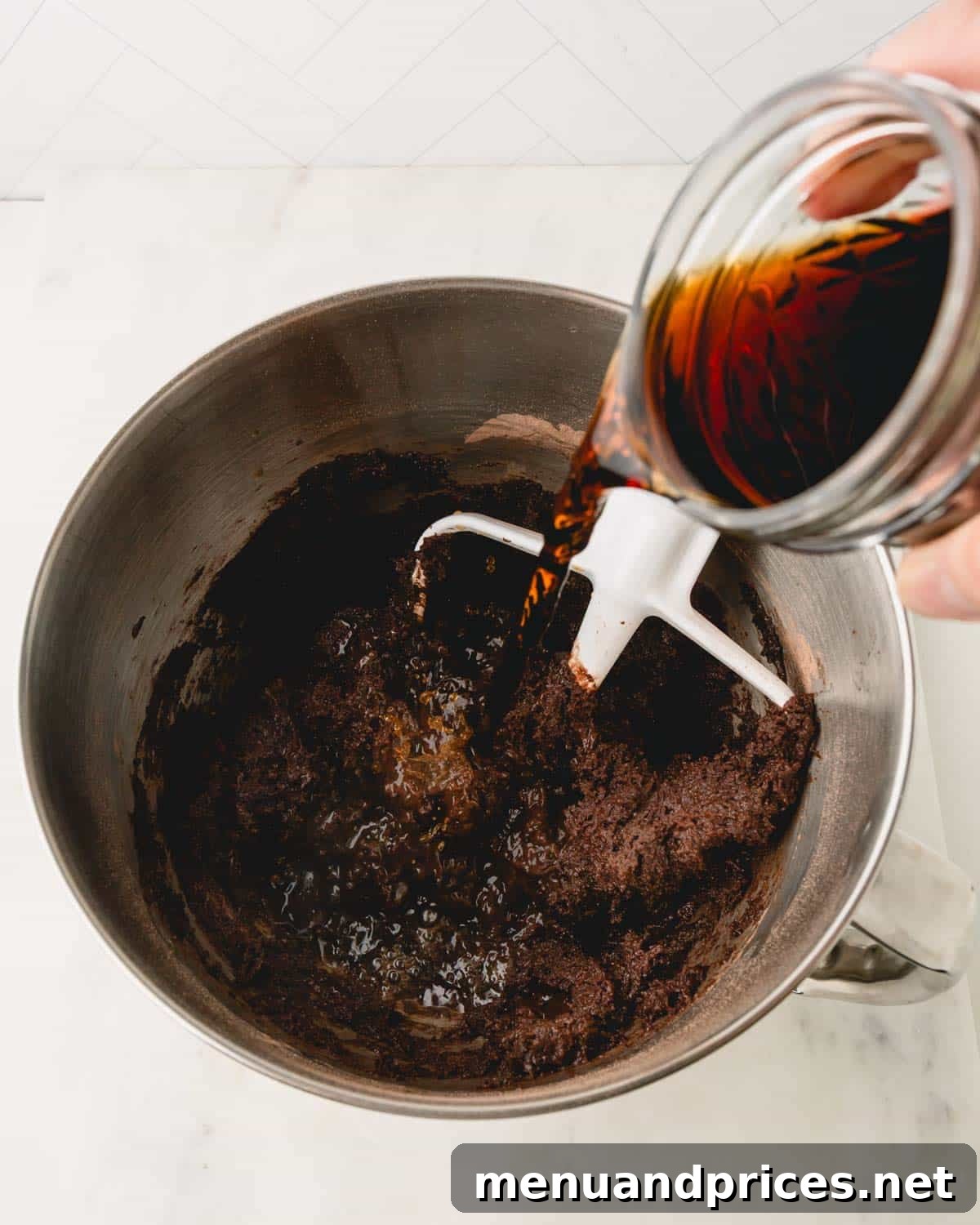 A hand pouring hot coffee from a glass jar into the German chocolate cake batter in a metal mixing bowl, blending it into the chocolate mixture.