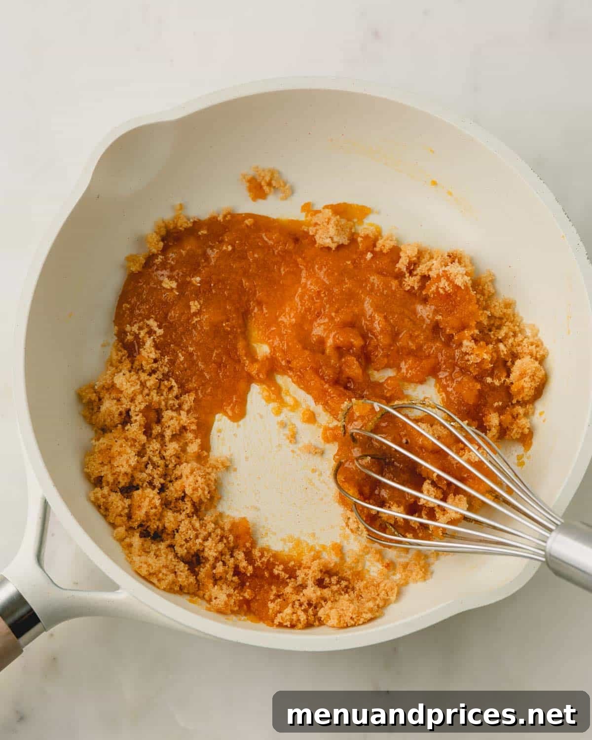 A white saucepan on a stovetop with brown sugar and egg yolk mixture, and a whisk resting on the side.