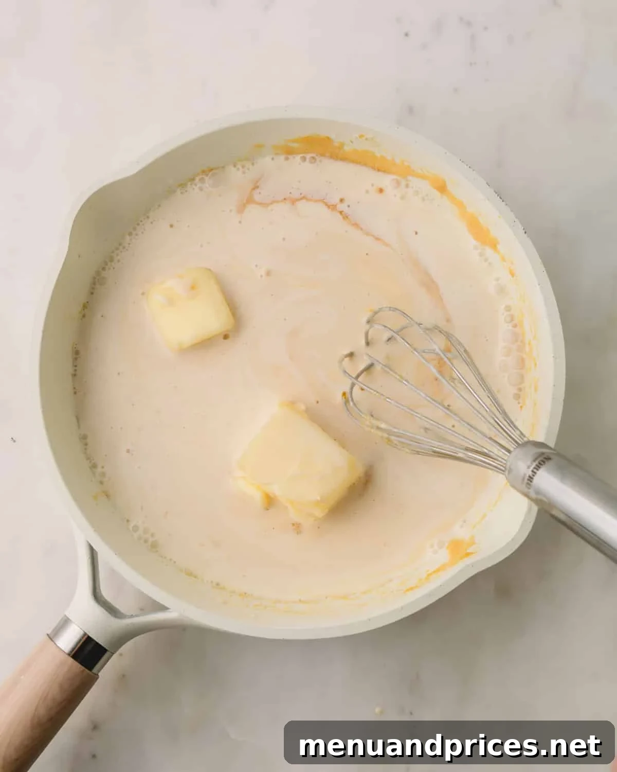 A saucepan on the stove containing a mixture of sugar, heavy cream, butter, and vanilla extract, with a whisk resting in it.