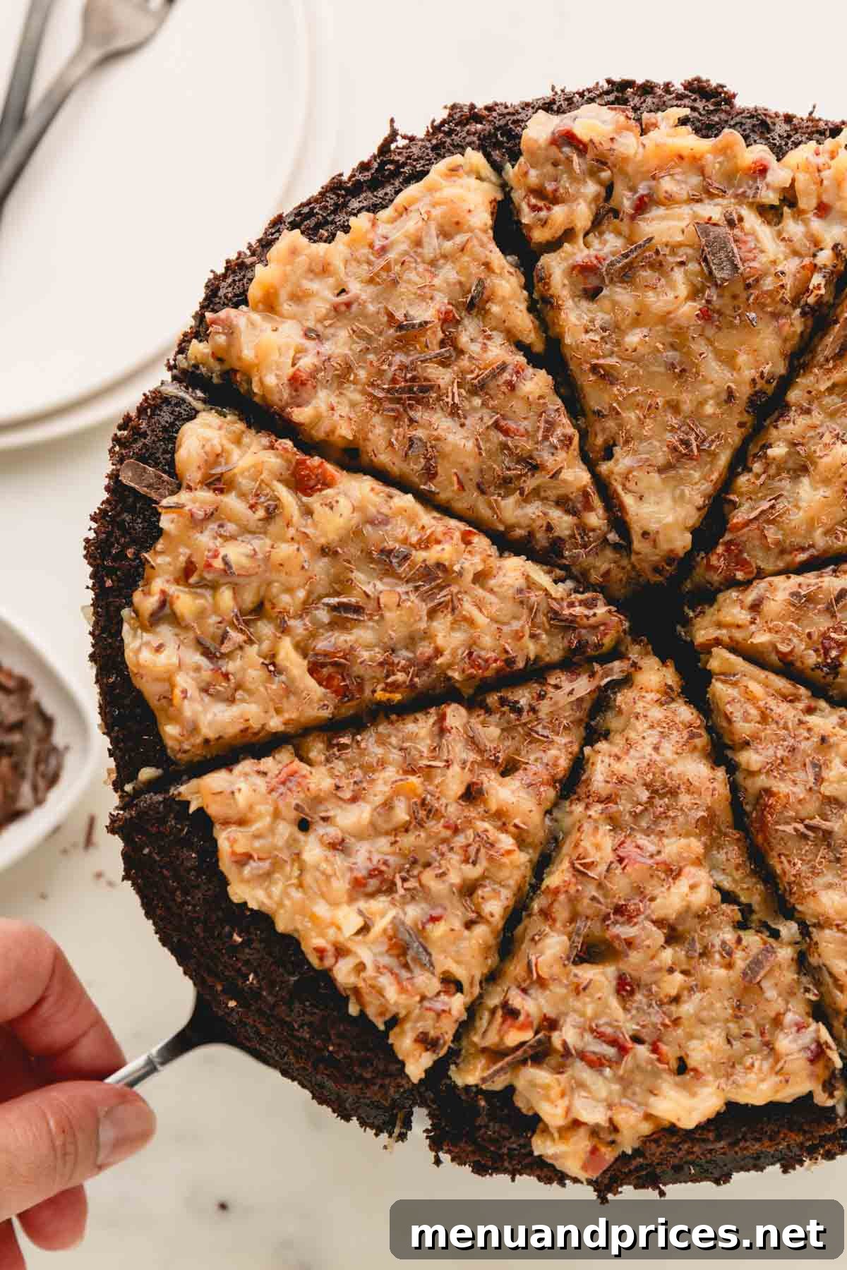A hand expertly taking a slice of three-layer German chocolate cake from a cake pedestal, highlighting the rich frosting between the moist chocolate layers.
