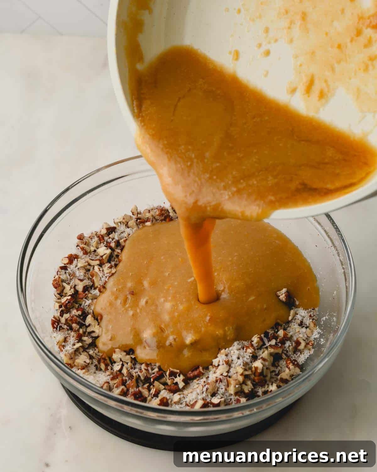 The melted brown sugar and butter mixture being poured from a saucepan over the coconut and pecans in a clear glass bowl, ready to be combined.