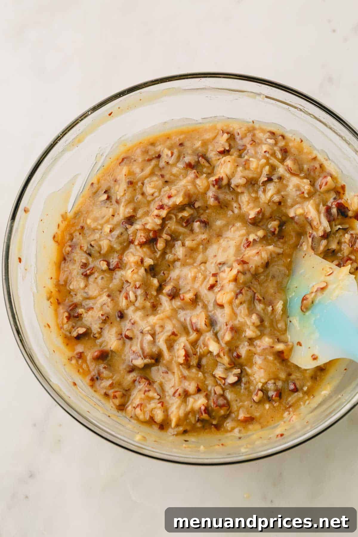 A glass bowl filled with the prepared coconut pecan frosting, showing its thick, textured consistency, with a spatula resting on the side.