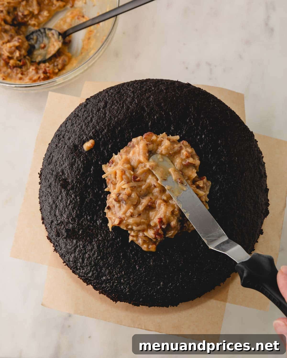 A German chocolate cake layer on a cake stand, with coconut pecan frosting being carefully spread over its surface.