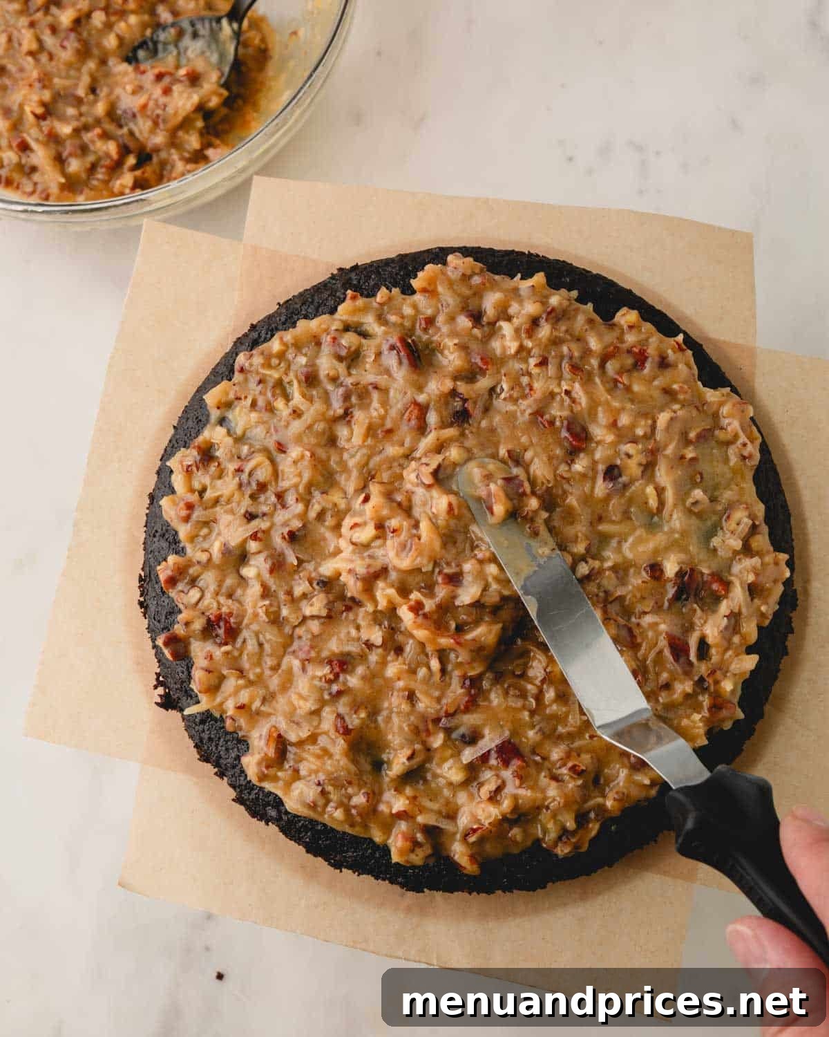 A hand spreading the coconut pecan frosting evenly over a layer of German chocolate cake, showing the rich texture of the frosting.