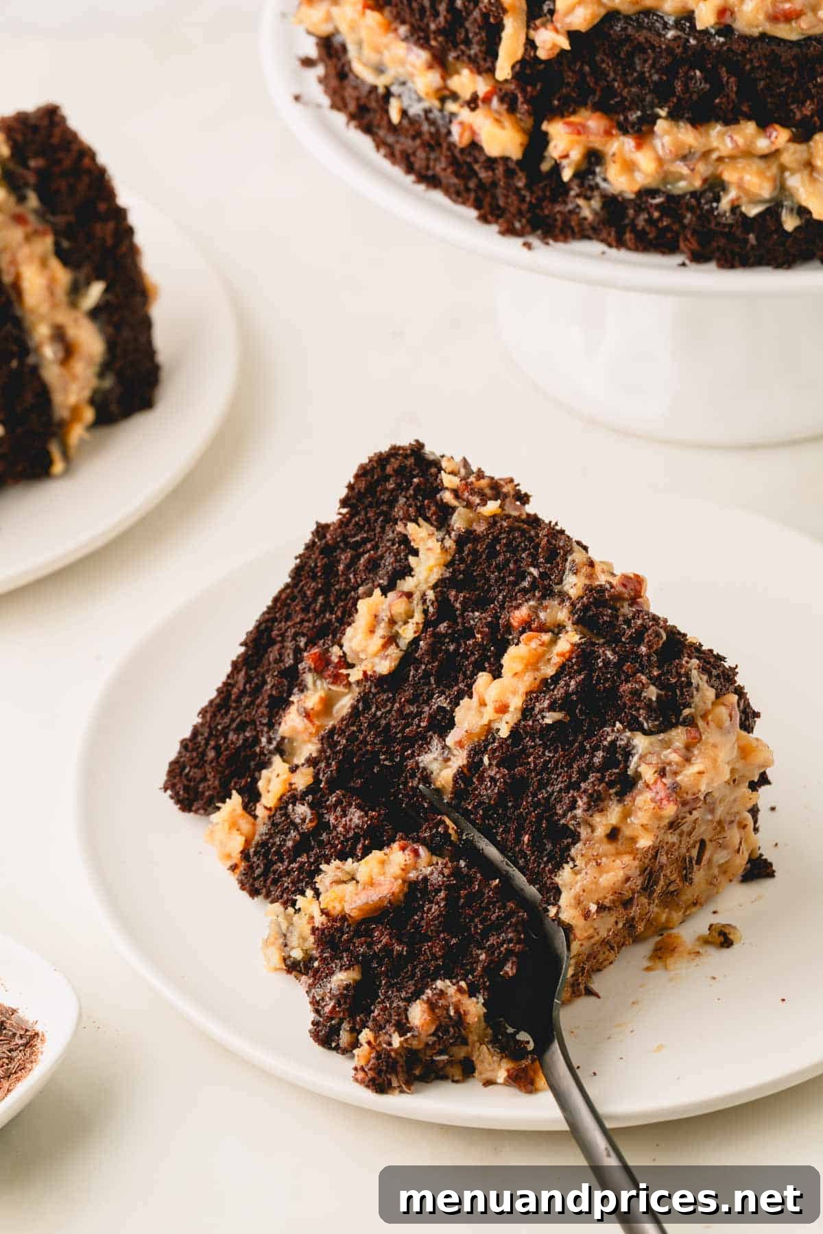 A white plate holding a single, perfectly cut slice of German chocolate cake, with a multi-layered cake on a pedestal in the soft background.