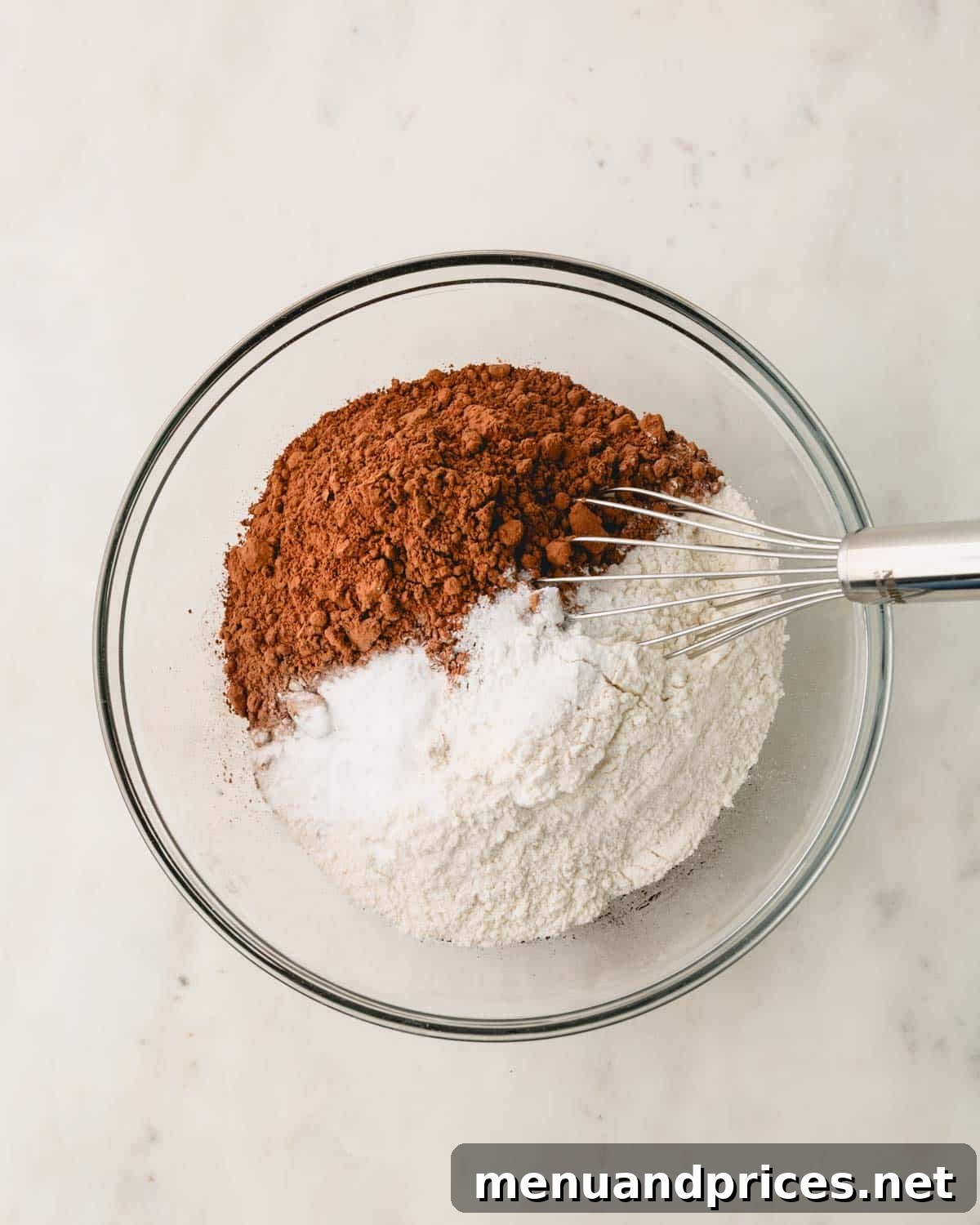A clear glass bowl containing all the dry ingredients for German chocolate cake, including flour, sugar, and cocoa powder.