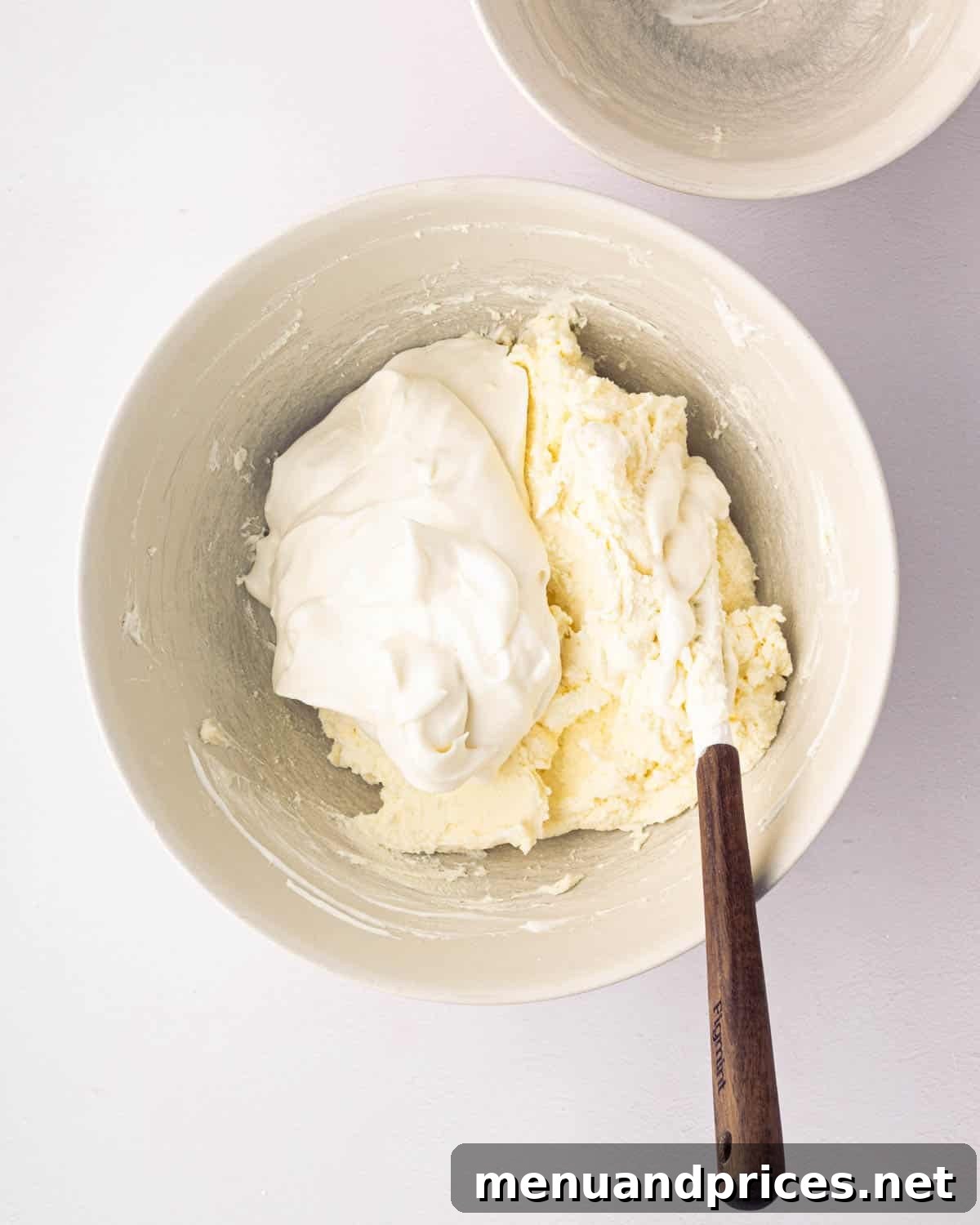 Bright Citrus Tiramisu 13 Whipped cream and mascarpone cheese being gently folded together with a wooden spatula in a white bowl.