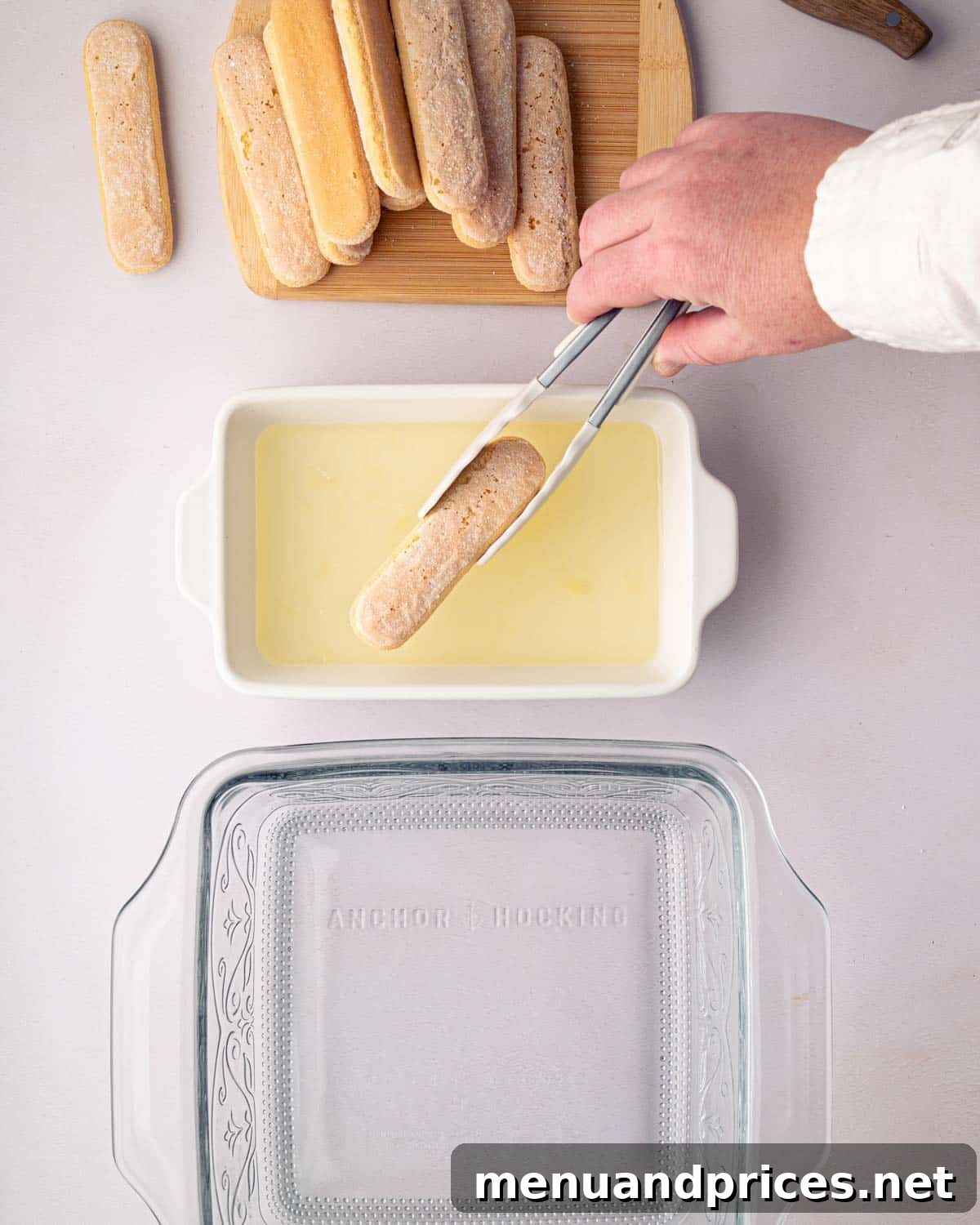 Bright Citrus Tiramisu 15 A hand dipping a ladyfinger into a bowl of limoncello simple syrup, with a glass baking dish waiting to be layered.