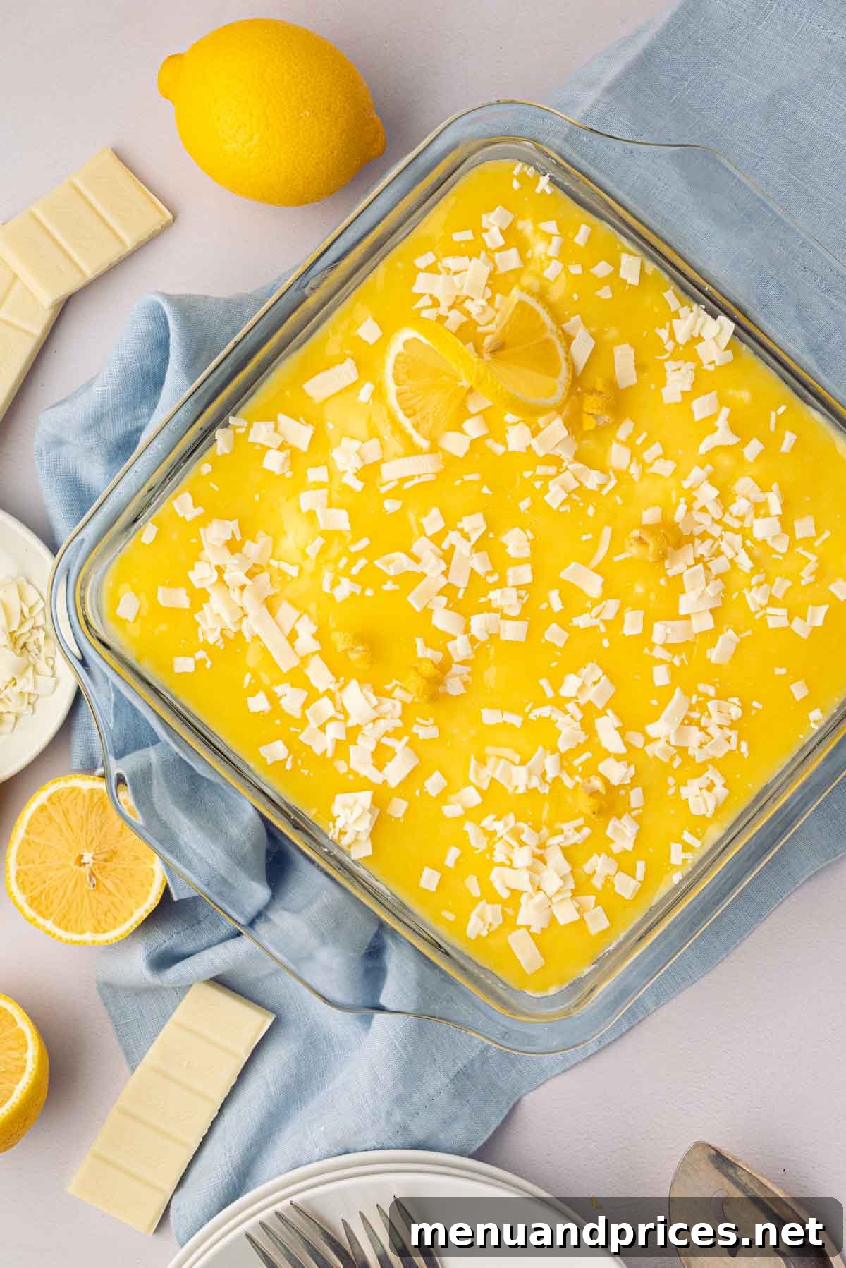 Bright Citrus Tiramisu 23 The fully assembled lemon tiramisu, garnished with delicate white chocolate shavings, positioned on a blue linen with fresh lemons for decoration, showcasing its vibrant appeal.