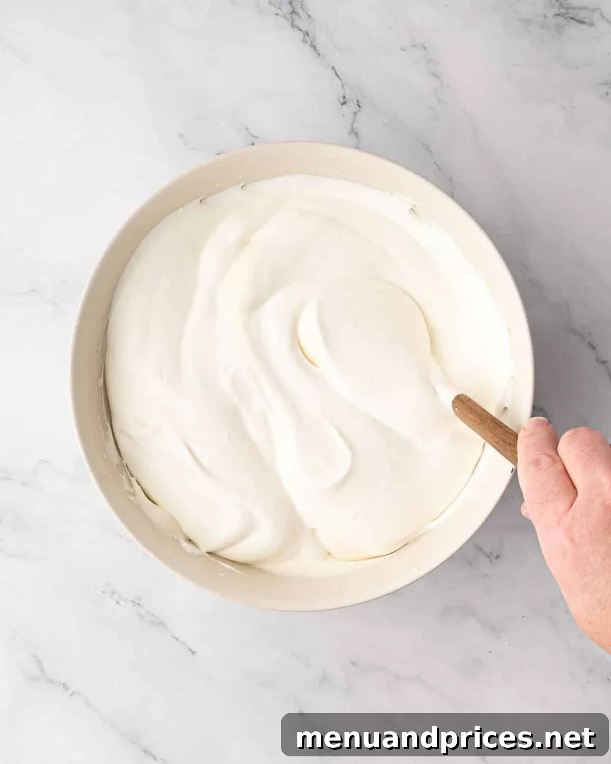 Whipped cream being gently folded into the mascarpone mixture with a spatula in a white bowl.