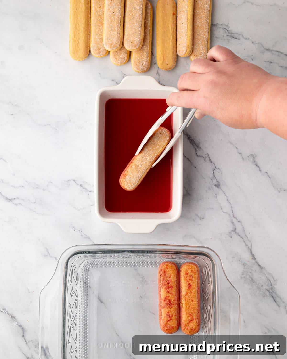 A hand dipping ladyfingers into raspberry simple syrup and carefully placing them into a glass baking dish.