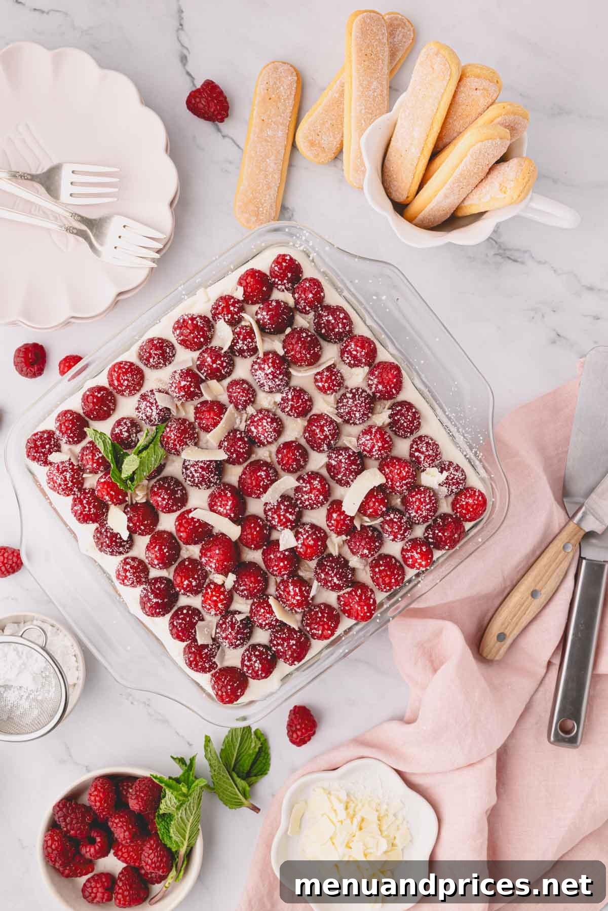 Finished raspberry tiramisu, beautifully garnished with fresh raspberries, ready to be served with plates nearby.