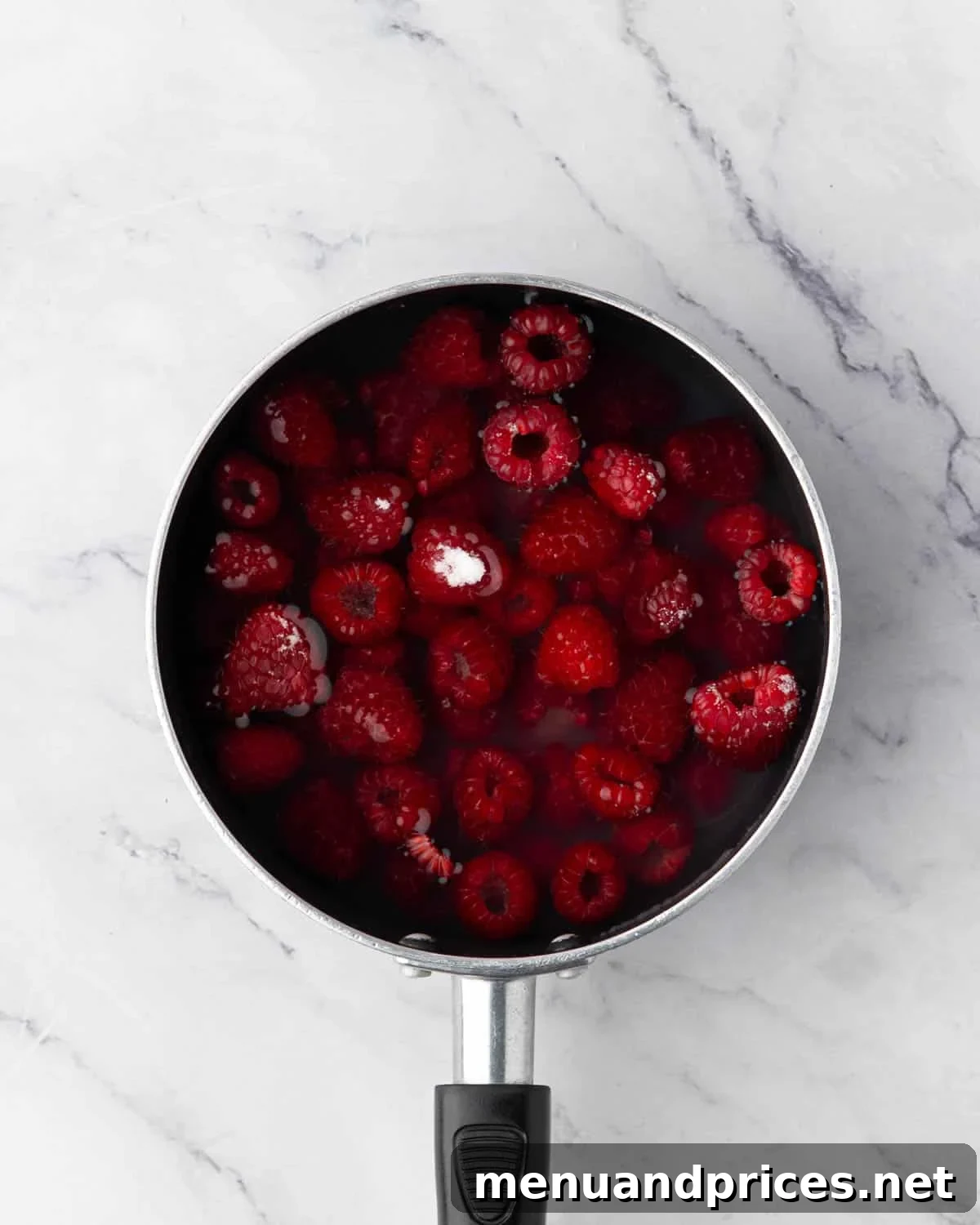 Fresh raspberries, sugar, water, and lemon juice in a black saucepan, ready to be cooked into syrup.
