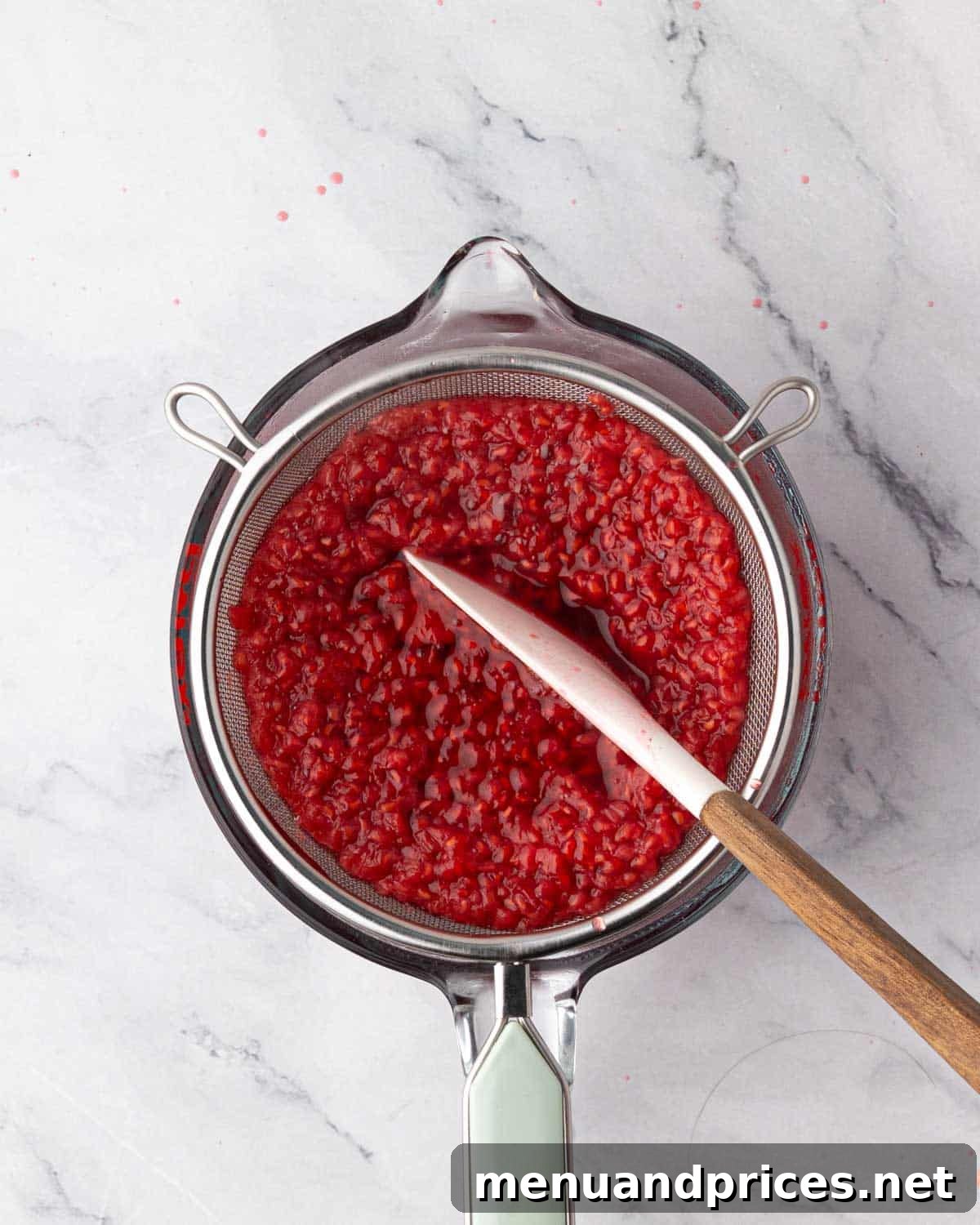 Straining raspberry simple syrup through a fine-mesh sieve into a glass measuring cup, with a spatula pressing the pulp.