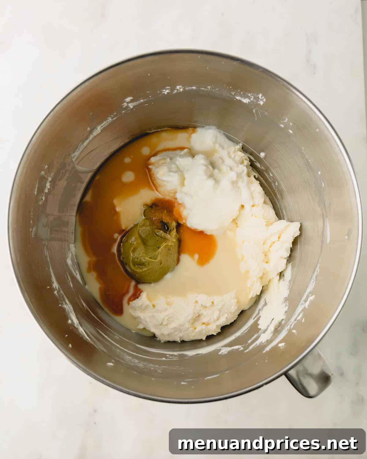 A stand mixer bowl with pistachio cheesecake filling ingredients inside. Pistachio butter and vanilla extract are on top.