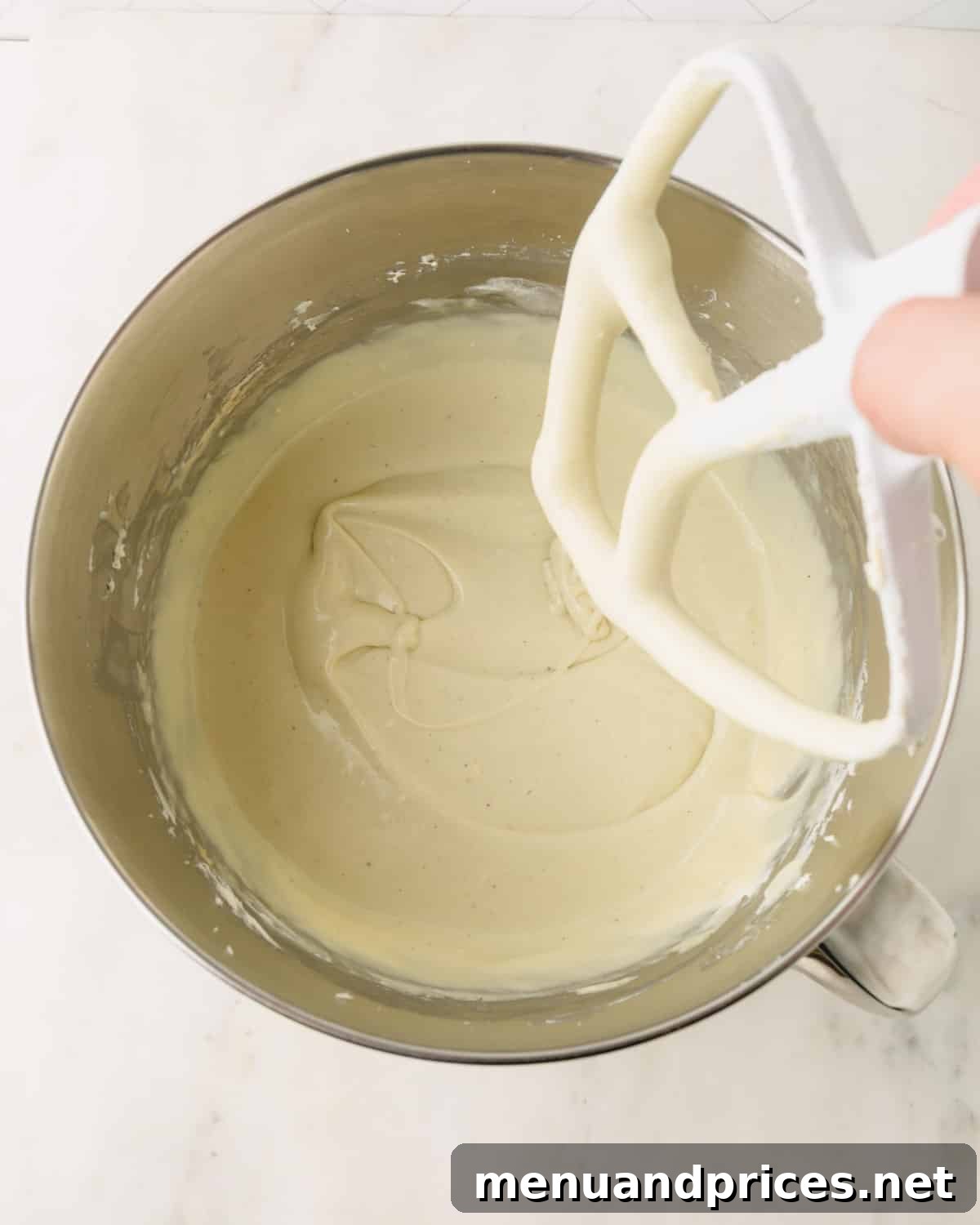 Pistachio cheesecake batter is combined in a stand mixer bowl with a hand removing the paddle attachment.