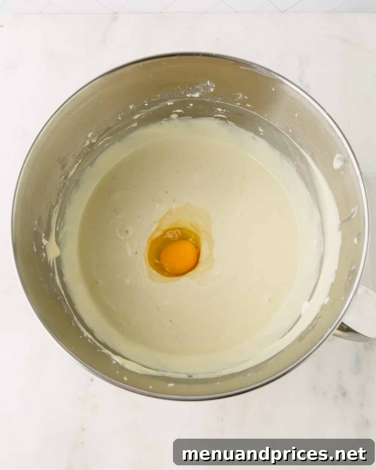 An egg is added to the pistachio cheesecake filling in a stand mixer bowl.