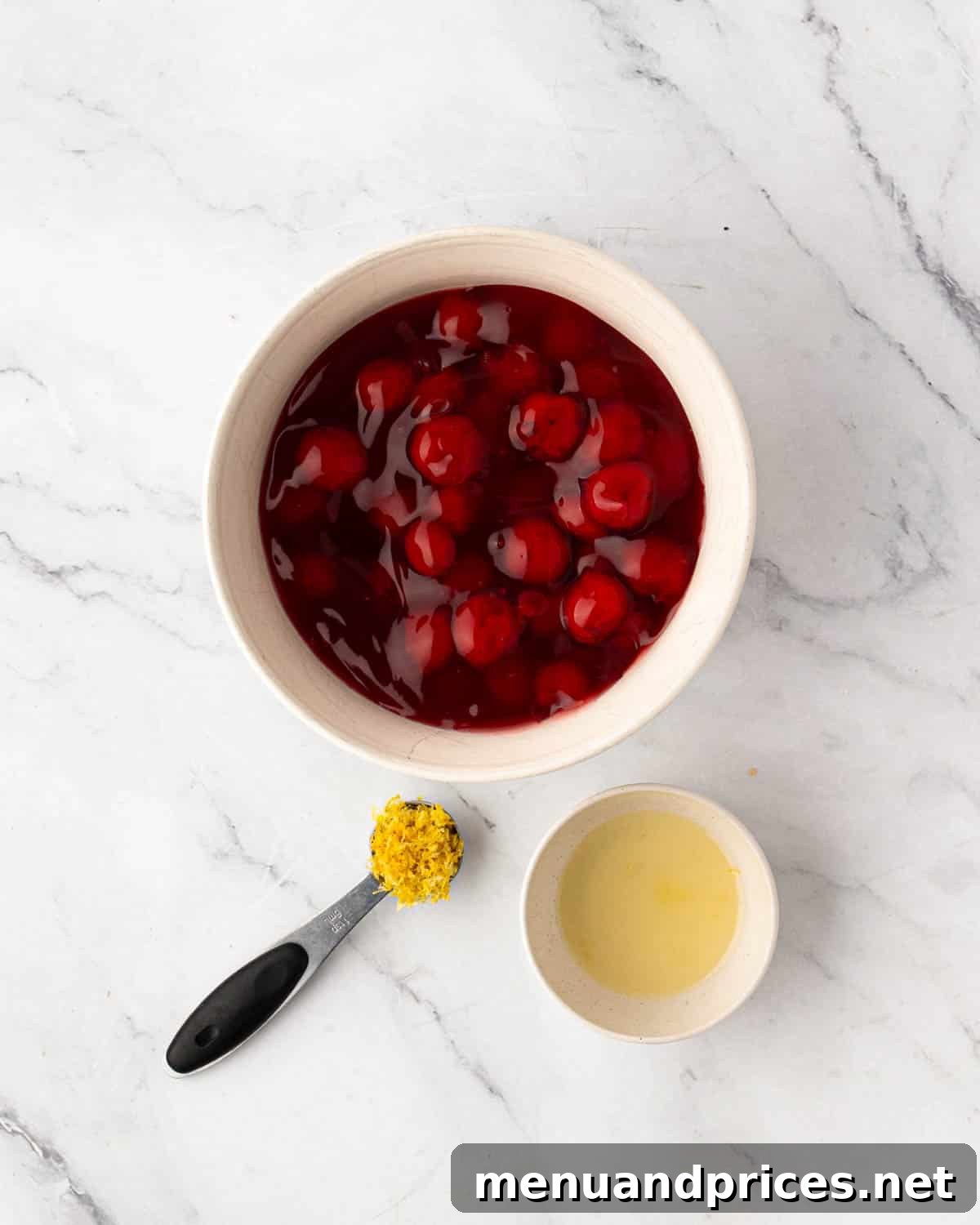 A white bowl with cherry pie filling, a measuring spoon with lemon zest, and lemon juice in a small white bowl.
